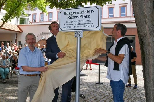 Bürgermeister Christoph Oeldorf (Mitte) enthüllt mit den beiden Enkeln Georg Rufers, Thomas (l.) und Matthias Rufer, das neue Platzschild. Foto: Dorn